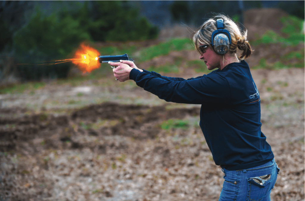 A woman firing a gun in a field wearing earmuff hearing protection