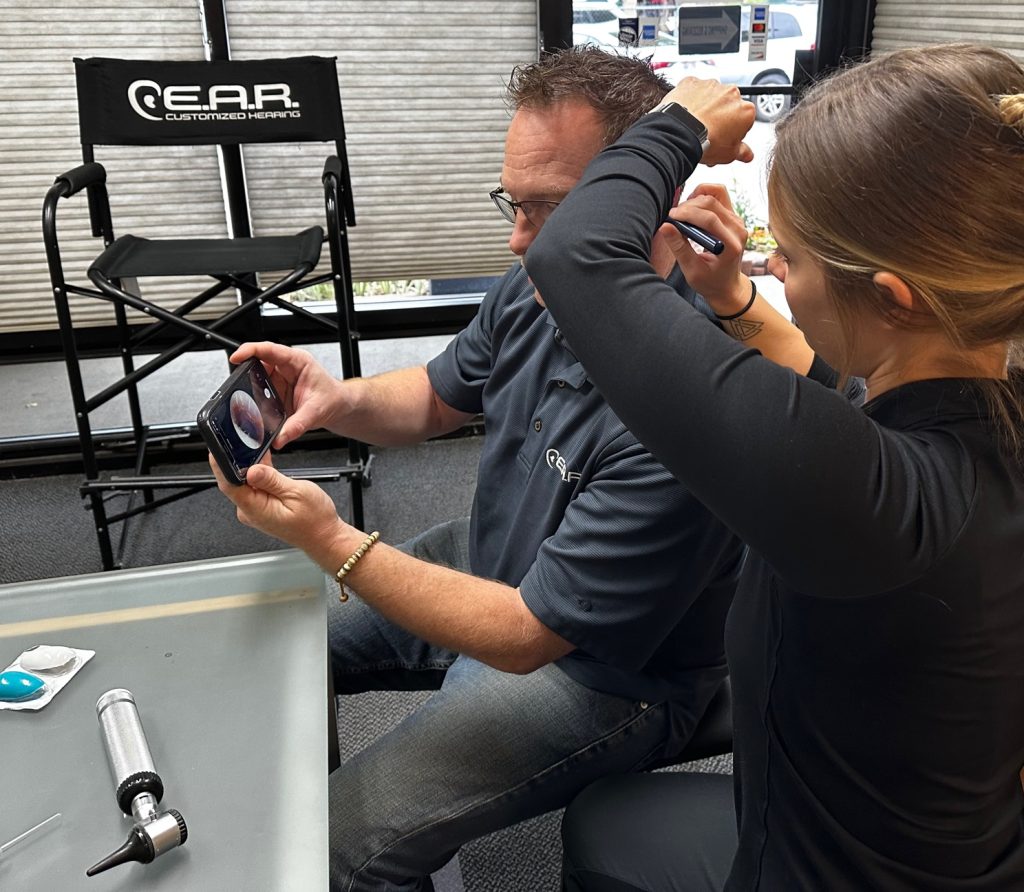 Man holds smartphone that displays his ear canal while a woman uses a digital otoscope.