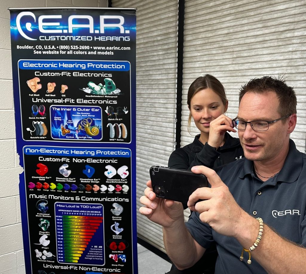 Woman examining a man's ear canal while he watches on a smartphone. Poster to the left of them describing custom hearing protection by E.A.R. Customized Hearing.